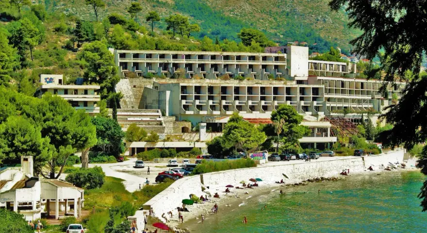 Opuszczone hotele Chorwacja, zniszczone budynki hotelowe na wzgórzu, plaża z ludźmi i morze.