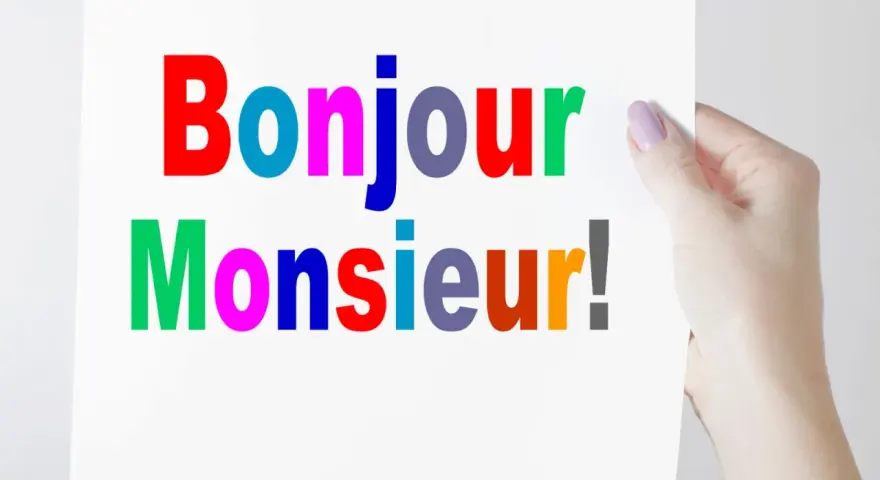 Una mano sostiene un cartel con "Bonjour Monsieur!" escrito en letras de colores.