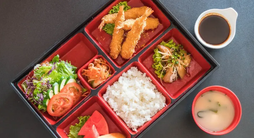 Japoński obiad w bento: smażone krewetki, ryż, sałatka, owoce i zupa miso.