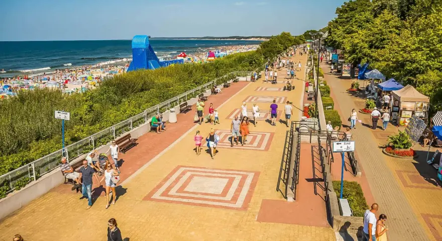 Tętniąca życiem ustka promenada w słoneczny dzień. Ludzie spacerują, odpoczywają na ławkach, a w tle widać plażę pełną turystów i morze.