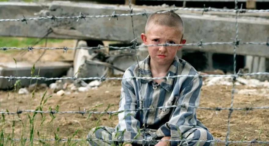 Pojken i randig pyjamas, Shmuel, sitter bakom taggtråd. Hans ansikte är smutsigt och hans blick är sorgsen.