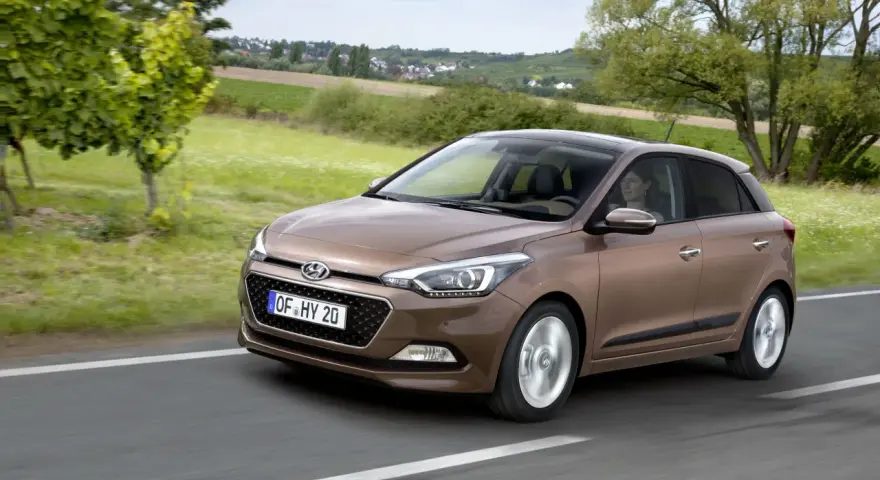 Brązowy Hyundai i20 na drodze wśród zieleni. Kierowca uśmiecha się, co może sugerować pozytywne hyundai i20 opinie.