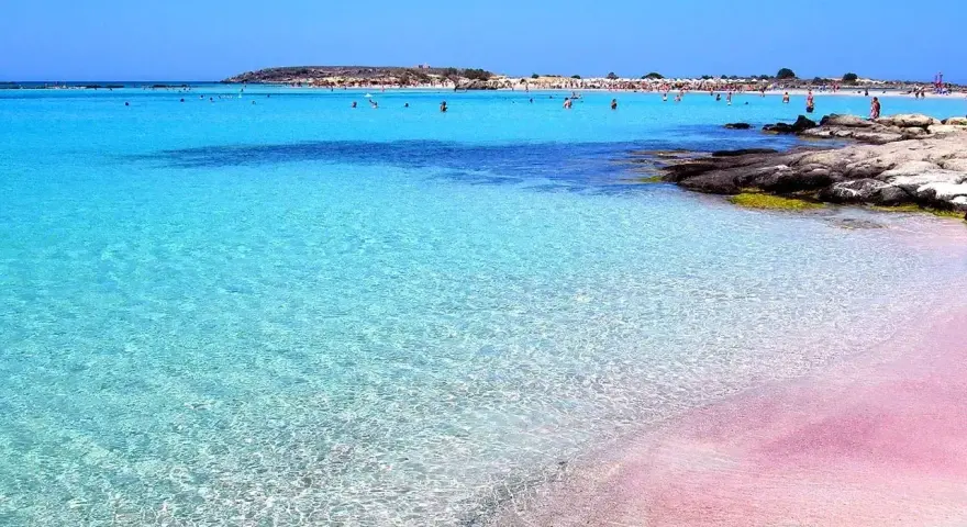 Różowy piasek i turkusowa woda na plaży Elafonisi. Ludzie kąpią się w krystalicznie czystym morzu.