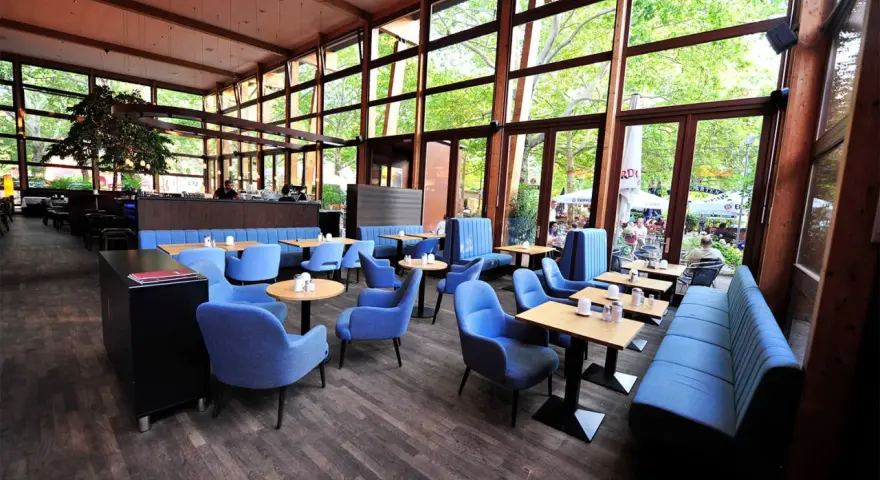 Gemütliches **Cafe Wilmersdorf** mit blauen Sesseln und Bänken, Holzboden und großen Fenstern, die auf eine grüne Terrasse blicken.