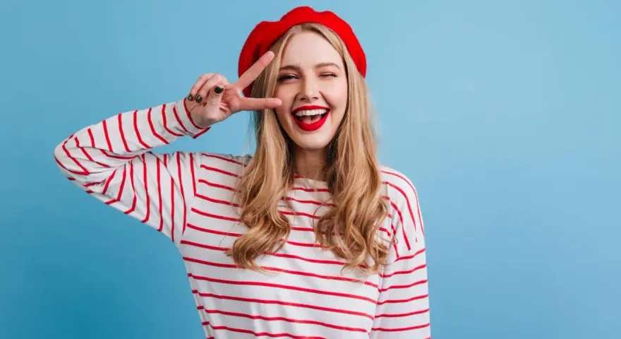 Mujer con boina roja y camiseta a rayas, haciendo el signo de la paz, con un estilo que evoca un caracter francés.