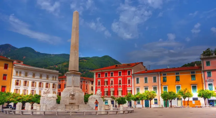 Piazza colorata con obelisco e leoni, incorniciata da edifici vivaci e montagne verdi, nella provincia di Massa e Carrara.
