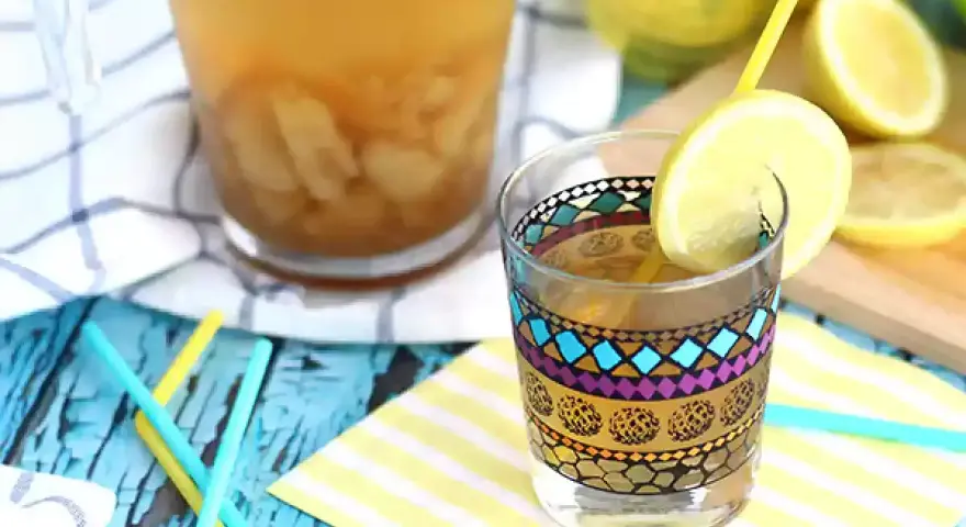 Ett glas med en färgglad dekorativ kant, fyllt med iste och en citronskiva. Bredvid står en karaff med iste och färska citroner. Perfekt iste recept för en varm dag.