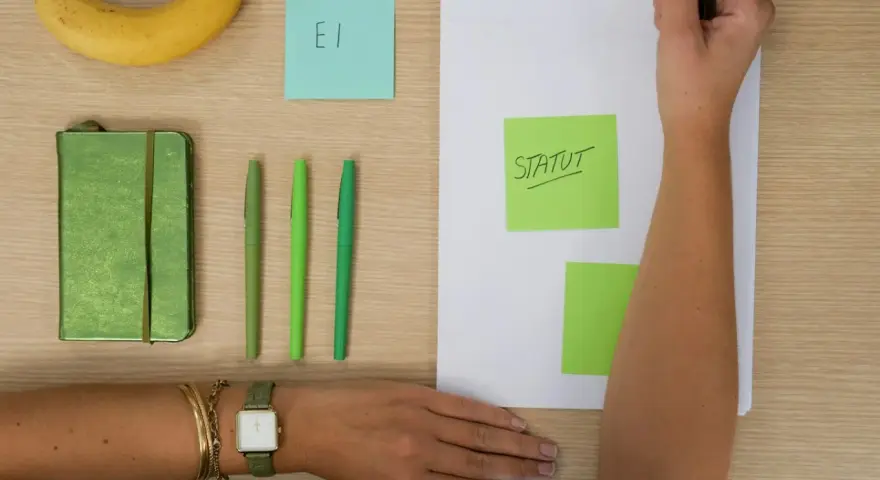 Une main écrit "STATUT" sur un post-it vert, à côté d'un carnet, de stylos et d'une banane. L'acronyme "EI" sur un post-it bleu suggère une **entreprise individuelle**.