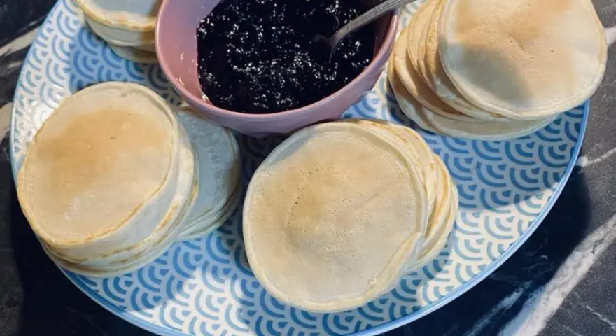 Une assiette bleue et blanche présente des piles de poffertjes dorés, accompagnés d'un bol de confiture de myrtilles. Parfait pour une recette de poffertjes !