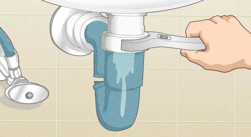 A hand uses a wrench to loosen a sink trap, a step in how to clean a sink drain. Water drains into a bucket below.
