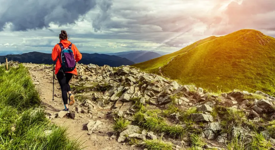 Kobieta w pomarańczowej kurtce wyrusza na szlak. To prawdziwy hiking po polsku, z pięknymi widokami i kamienistą ścieżką.