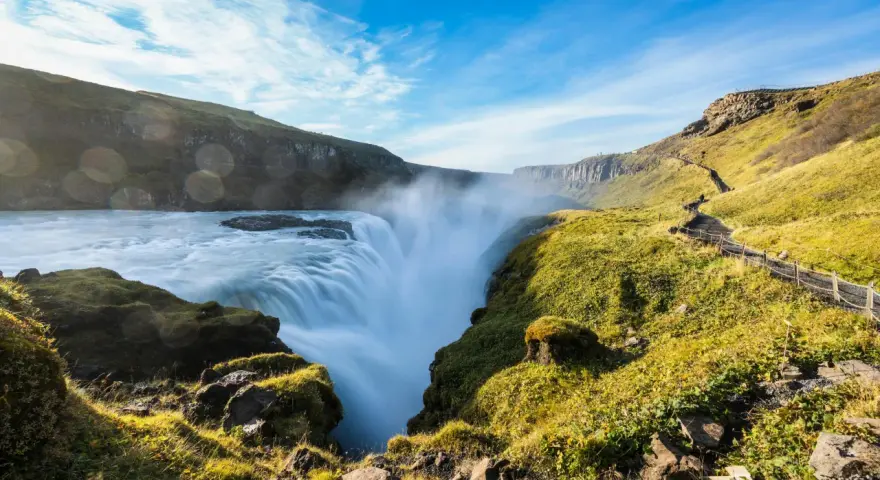 Potężny wodospad Gullfoss w Islandii, z mgiełką unoszącą się nad kaskadami. Ścieżka turystyczna wije się po zielonym zboczu.