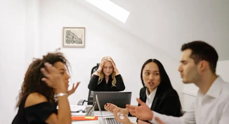 Un gruppo di colleghi di lavoro discute animatamente. Una donna bionda sembra stressata, tenendosi la testa tra le mani.