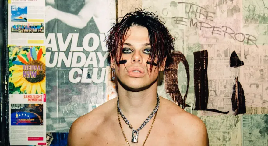 Yungblud, "The Emperor", z charakterystycznym makijażem i łańcuchami, na tle plakatów.