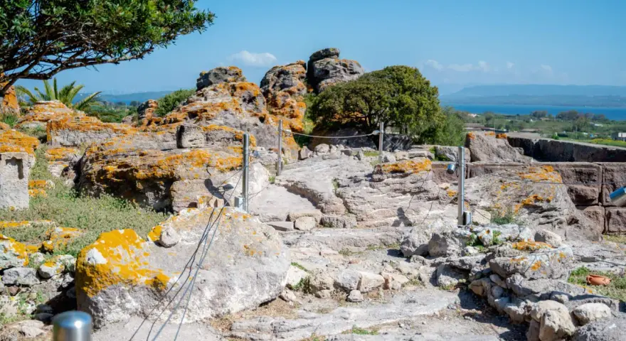 Ancient Italian ruins with weathered stones covered in orange lichen, a pathway winding through the site, and a distant blue sea under a clear sky.