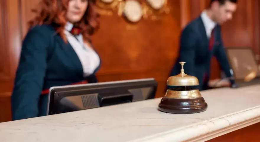 Recepcjonistka i recepcjonista za ladą hotelową. Dzwonek do obsługi gości, który pomoże dowiedzieć się, ile kosztuje doba hotelowa.