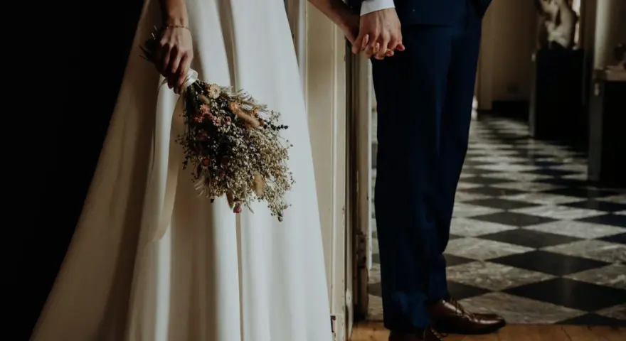 Para młoda trzyma się za ręce, ona z bukietem suszonych kwiatów. Wesele motywy w eleganckim wnętrzu.