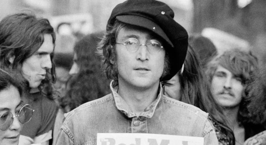 John Lennon w czapce i okularach, trzymający plakat "Red Mole". W tle inni ludzie, być może fani jego utworów.
