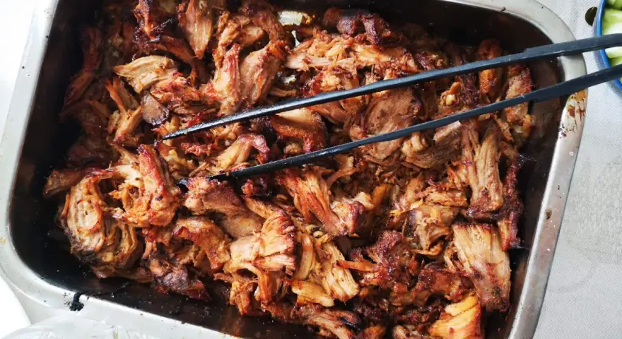 Saftigt, långkokt fläskkött i en ugnsform, perfekt för ett carnitas recept. Tång ligger redo att servera.