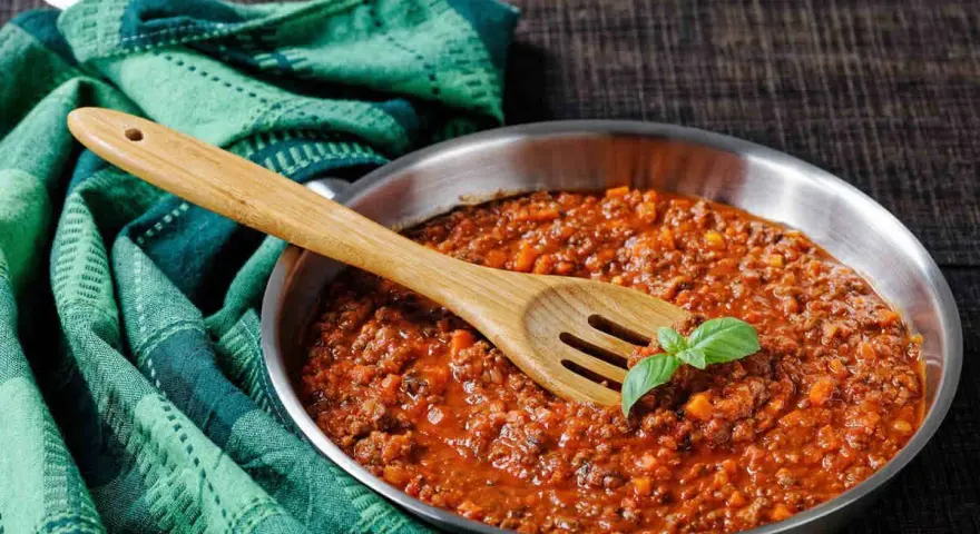 Un saporito ragù di cinghiale in padella, pronto per condire la pasta. Un cucchiaio di legno e una fogliolina di basilico completano la scena.