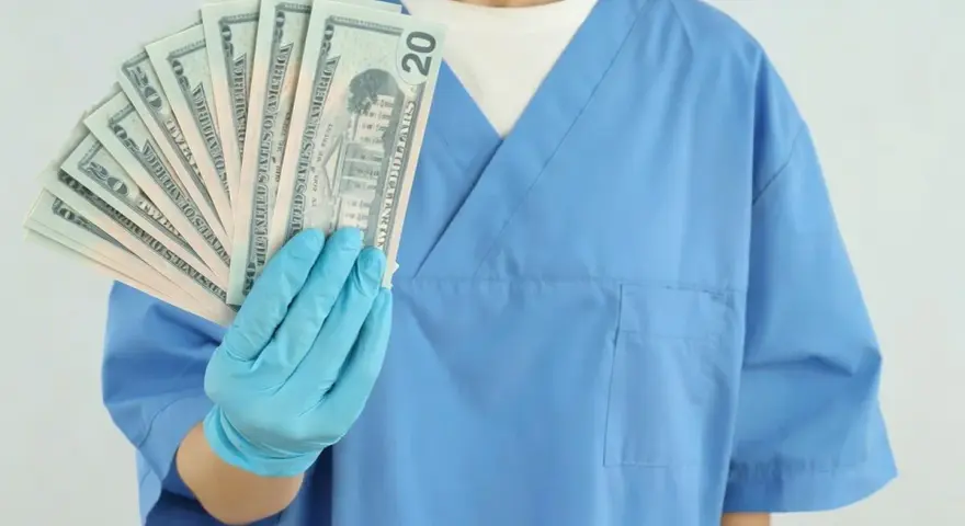 Osoba w niebieskim fartuchu i rękawiczkach trzyma wachlarz banknotów 20 dolarowych.