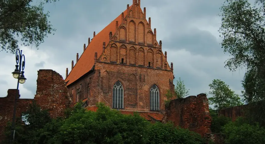 Gotycka ceglana budowla z pomarańczowym dachem, otoczona zielenią i fragmentami murów. Widok na Elbląg, województwo warmińsko-mazurskie.