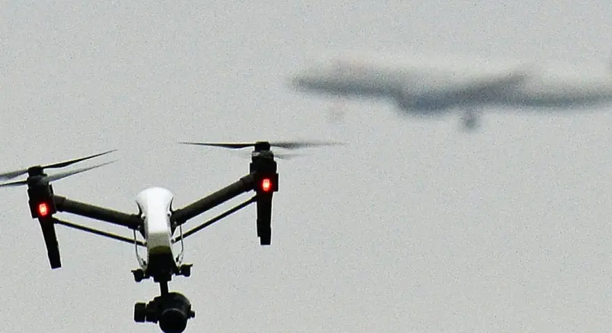 A white drone with red lights hovers in the sky, possibly on a security mission, as a blurred airplane flies in the distance.