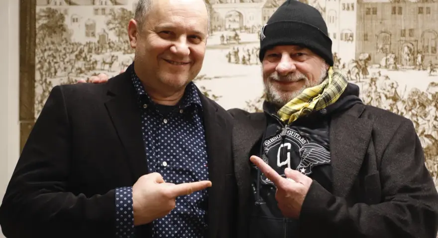 Deux hommes souriants, l'un avec une chemise à pois, l'autre avec un bonnet, se montrent du doigt. Un chanteur de confiance.