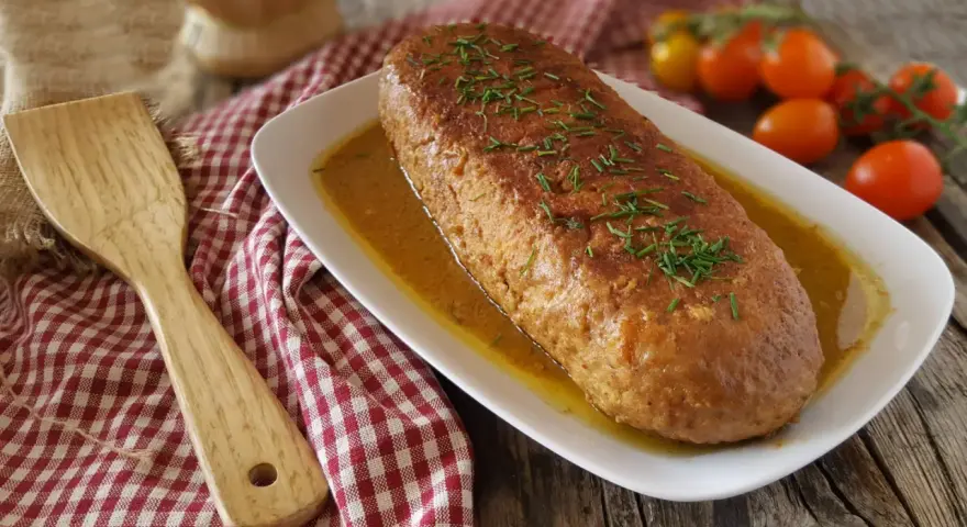 Polpettone in padella dorato, guarnito con erba cipollina, servito su un piatto bianco con salsa. Accanto, una spatola di legno e pomodorini.