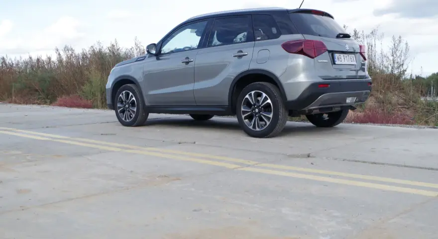 Szary Suzuki Vitara Hybrid z czarnym dachem stoi na betonowej nawierzchni z żółtymi pasami.