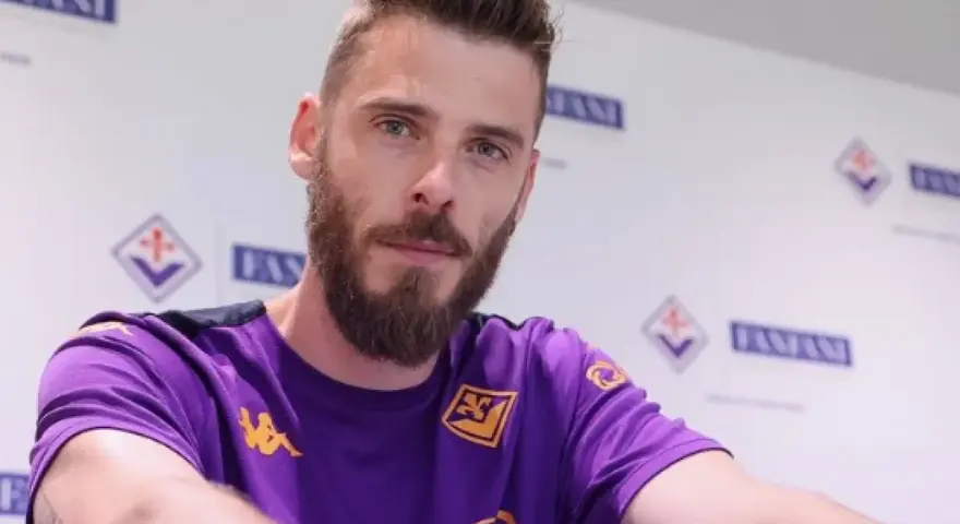 David de Gea, con barba y camiseta morada, posa ante un fondo con logos.
