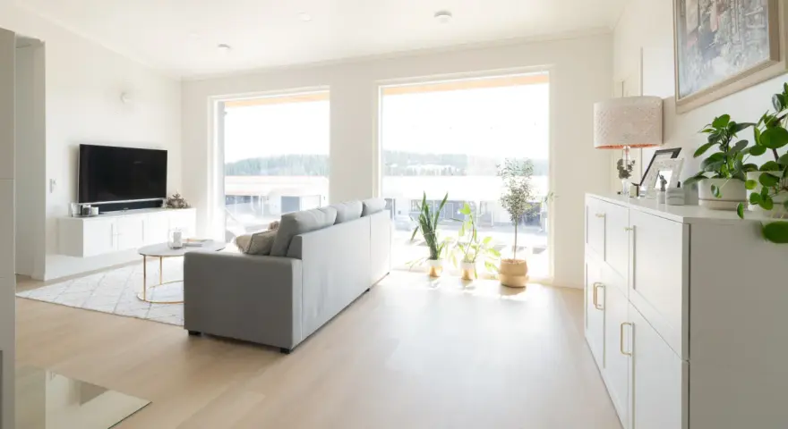 Ett ljust vardagsrum med en grå soffa, ett runt soffbord och en vit förvaringsmöbel. En lampa med vitt ljus lyser upp rummet.