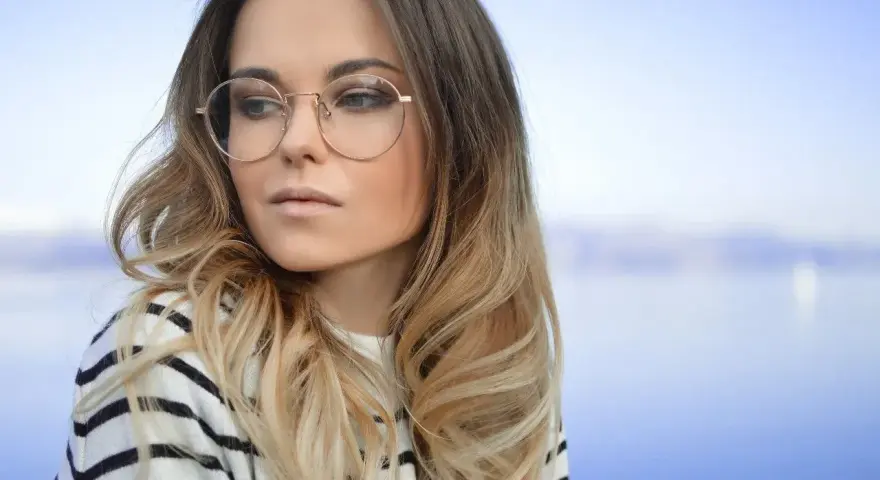 Blondynka w okularach o okrągłych oprawkach, w bluzie w paski, patrzy w bok na tle błękitnego nieba i wody.