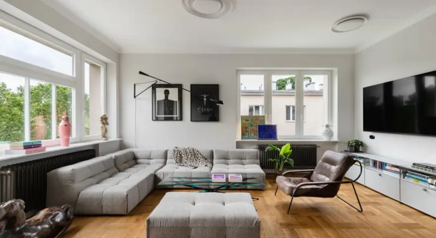 Nowoczesny salon z szarym narożnikiem, szklanym stolikiem i brązowym fotelem. Widać dzieła sztuki, rośliny i telewizor. To przykład udanego **retrofit** mieszkania.