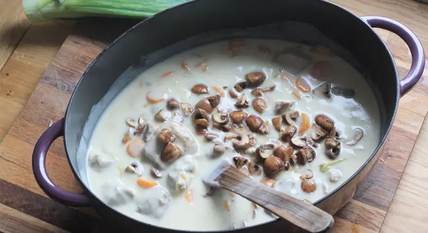 Un plat de blanquette de veau facile mijote dans une cocotte, garni de champignons et de carottes. Un poireau est posé à côté.