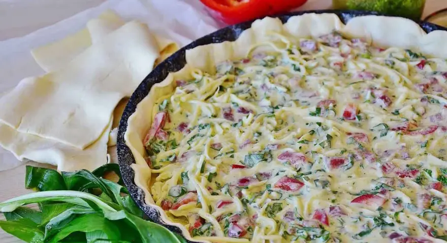 Tarte salée garnie de fromage, poivrons et ail des ours, prête à cuire. Pâte feuilletée et poivron rouge à côté.