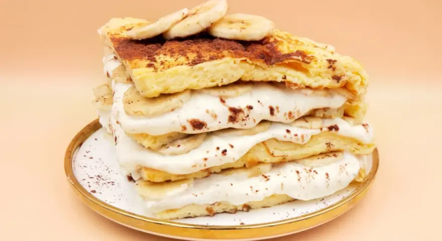Wysoki omlet z bananem, przełożony kremem i plasterkami banana, posypany kakao. Pyszny deser na talerzyku.