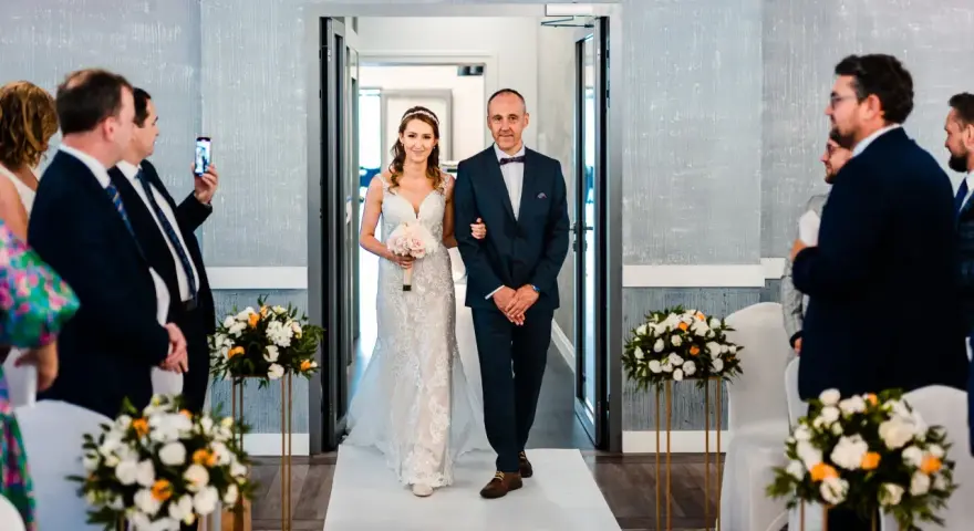 Panna młoda z ojcem idzie do ołtarza. W tle goście, a na stole bukiety kwiatów. Zastanawiasz się, jakie dokumenty do ślubu cywilnego?