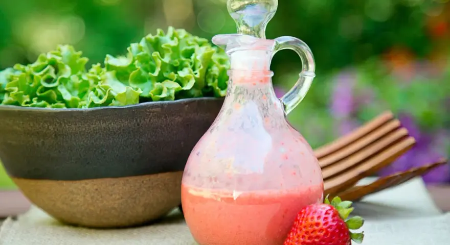 Erdbeeren mit Balsamico-Dressing in einer Karaffe, daneben eine Schüssel Salat und eine einzelne Erdbeere.