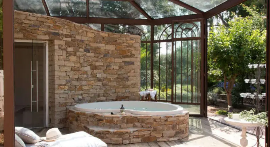 Un spa dans une cave, avec un jacuzzi en pierre, une banquette et une verrière donnant sur un jardin luxuriant.