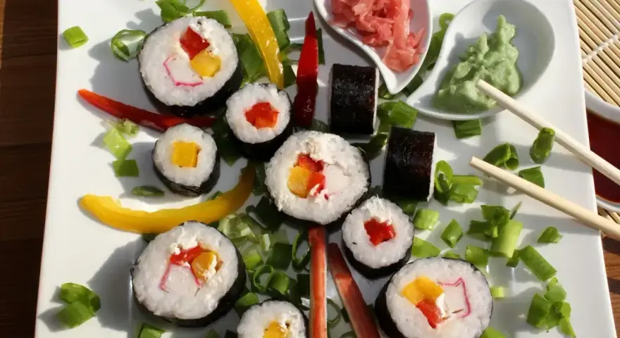 Kolorowe rolki sushi z surimi, papryką i mango, ozdobione szczypiorkiem i papryczkami chili. Obok wasabi i marynowany imbir.