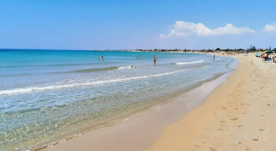 A wide, sandy beach with clear turquoise water and a few people swimming and sunbathing under a bright blue sky.