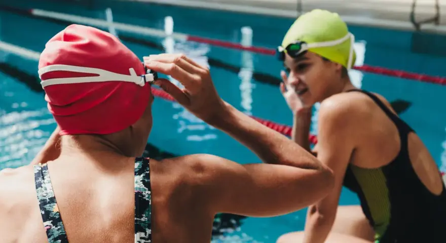 Deux nageuses se préparent pour une course de nage brasse. L'une ajuste ses lunettes, l'autre sourit.