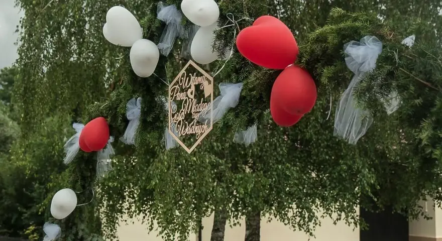 Drewniany napis "Witamy Parę Młodą" na gałęziach iglaków, ozdobionej białymi i czerwonymi balonami. Idealne śmieszne teksty na bramy weselne!
