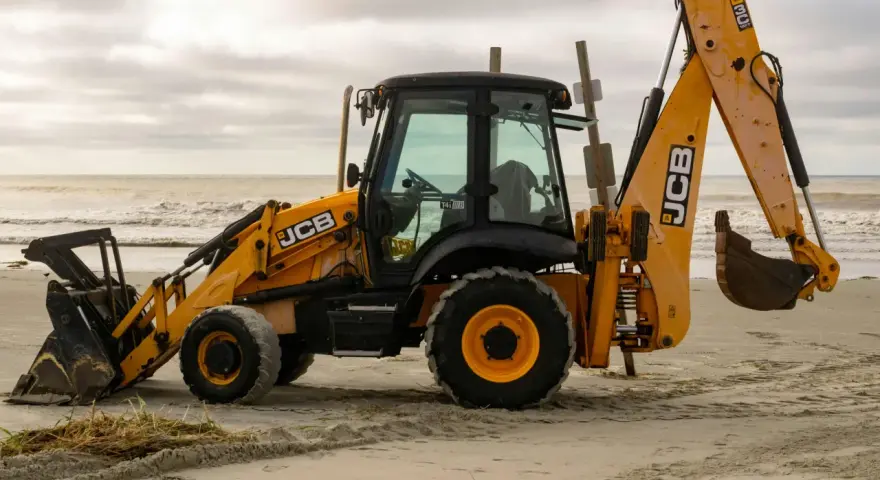 Żółta koparko-ładowarka firmy JCB stoi na piaszczystej plaży, gotowa do pracy.