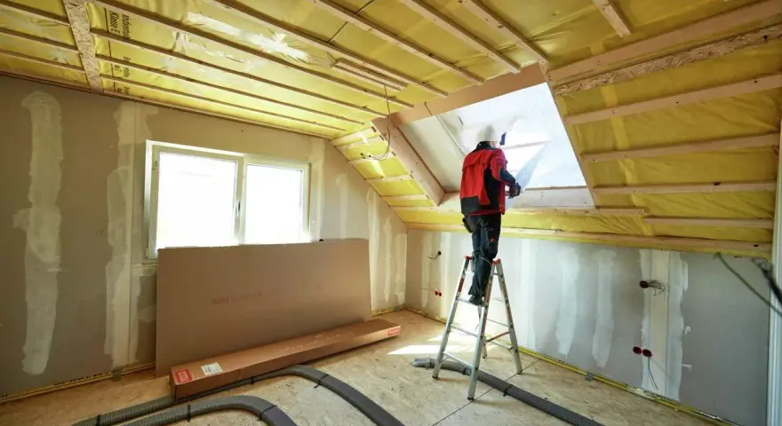 Operaio in piedi su una scala per isolare il tetto di una mansarda con materiale isolante giallo.