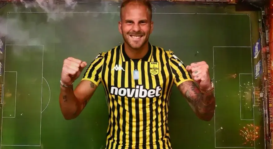 Uros Racic, con su nueva camiseta a rayas amarillas y negras, celebra con los puños en alto. El fondo simula un campo de fútbol con bengalas.