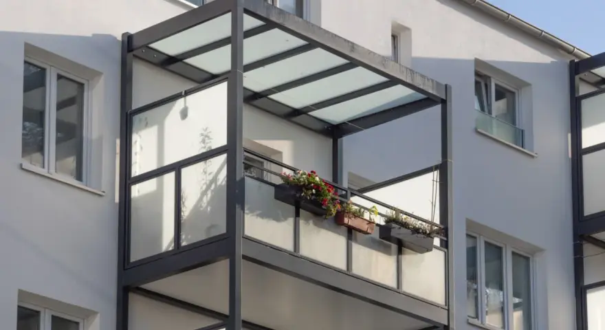 Nowoczesny balkon dostawny z przeszklonym zadaszeniem i balustradą, ozdobiony skrzynkami z kwiatami. Sprawdź balkon dostawny cena.