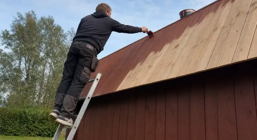 Man på en stege målar plank på ett tak med brun färg.