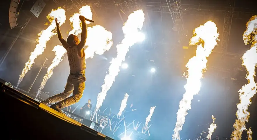 Un chanteur de groupe hard rock des années 2000, micro en main, saute sur scène entouré de jets de flammes spectaculaires.